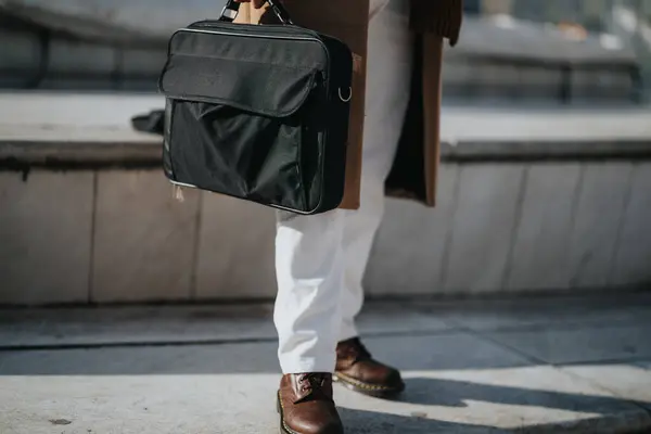 A lower body shot of a professional businessman in a stylish coat and white pants holding a laptop bag, walking confidently on an urban street.
