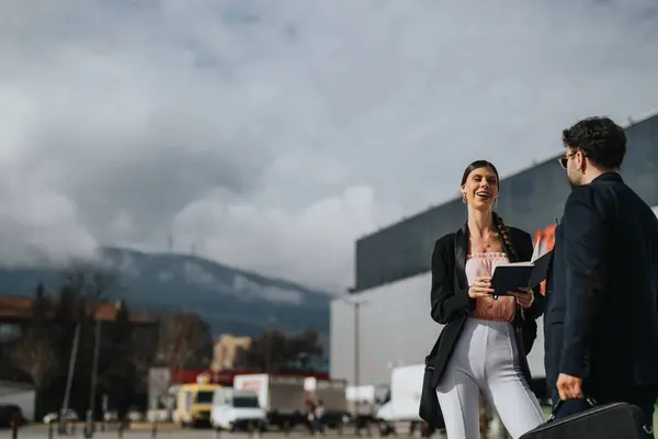 A man and woman in business attire laugh together during an informal outdoor meeting with a mountainous backdrop.