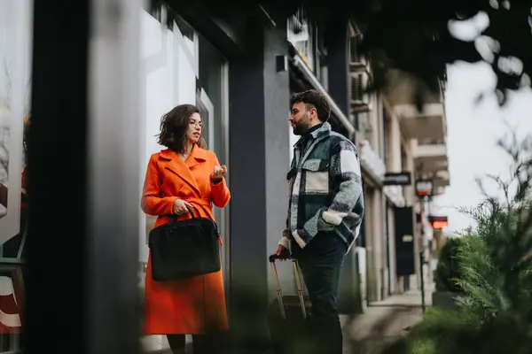 穿着鲜艳橙色外套的优雅女人与一位时尚男人交谈 两人都在城市人行道上提着行李 — 图库照片