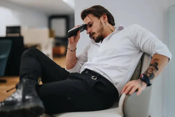 カジュアルなオフィス環境に座っている困惑した若い起業家は 額にスマートフォンを保持し 疲れているように見えます — ストック写真