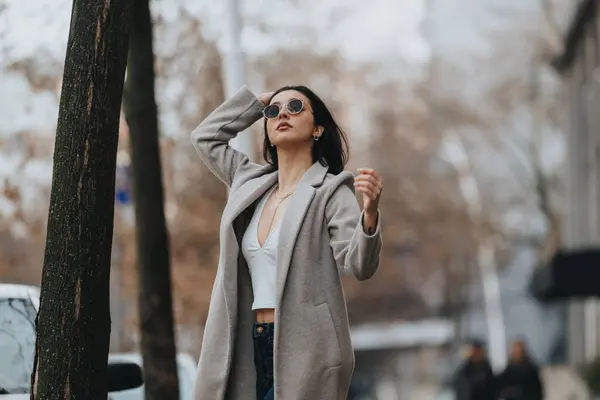 Stylish young woman in sunglasses and a chic coat poses outdoors on a city street, exuding elegance and autumn fashion vibes.