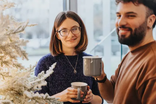 Smiling friends sharing a joyful moment with coffee by a Christmas tree.
