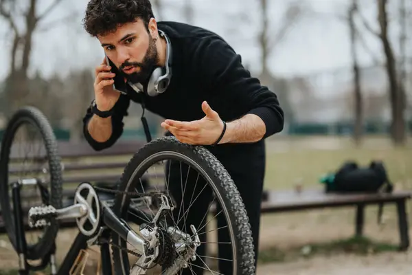 Focused businessman multitasking by fixing a bicycle in the park while talking on the phone, showing problem-solving skills.