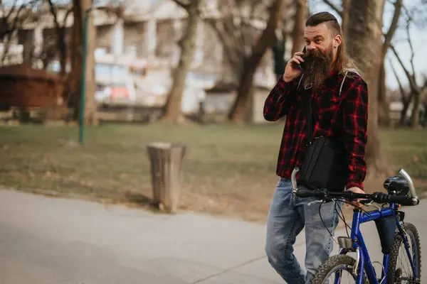 Urban professional with a beard making a business call while standing with his bicycle in a city park.