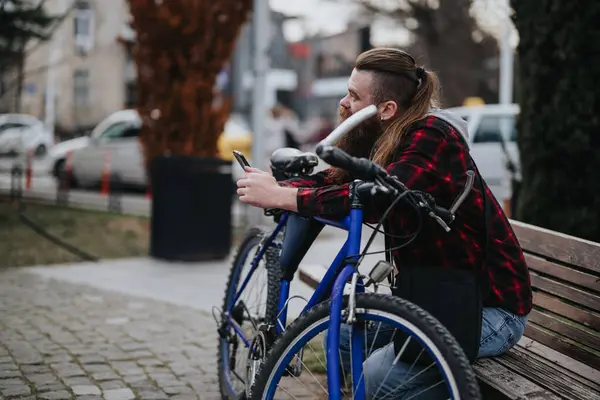 一个身穿休闲装 留着胡子的年轻人坐在城市长椅上 一边检查手机 — 图库照片