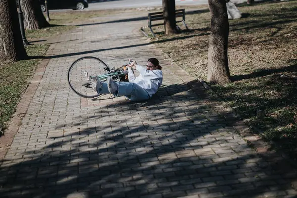 A cyclist has fallen off their bike onto a brick pathway in a sunlit park, capturing a moment of an unexpected accident.