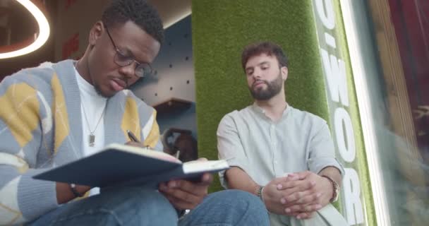 黒人の男性は 仕事で休憩中に閾値に座っている間 ノートブックに同僚を描いています — ストック動画