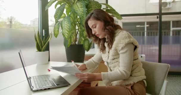 Ярком Офисе Молодая Женщина Сидит Концентрируется Написании Заметок Ноутбуком Рядом — стоковое видео