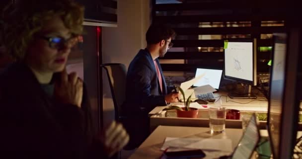 2人のビジネス同僚は 暗く照らされたオフィスで夜に仕事に深く魅了され プロのワークスペース内の献身とチームワークを例示しました — ストック動画