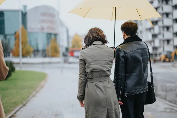 Back view of a romantic couple sharing a yellow umbrella, walking together on a wet sidewalk in an urban setting during autumn.