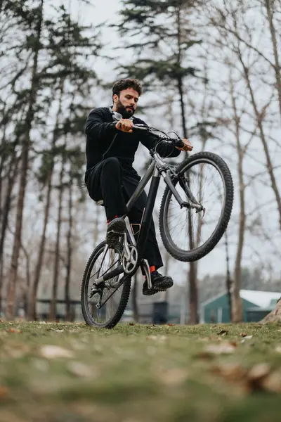 Focused man performing bicycle stunt in a forested park setting.