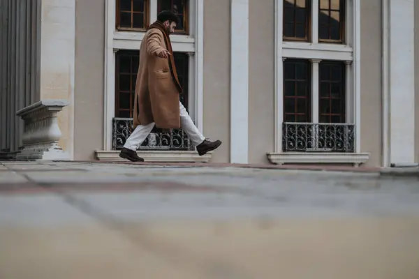 Fashionable male figure in a beige overcoat and dark pants strides past a classic architectural background, exuding confidence and sophistication.