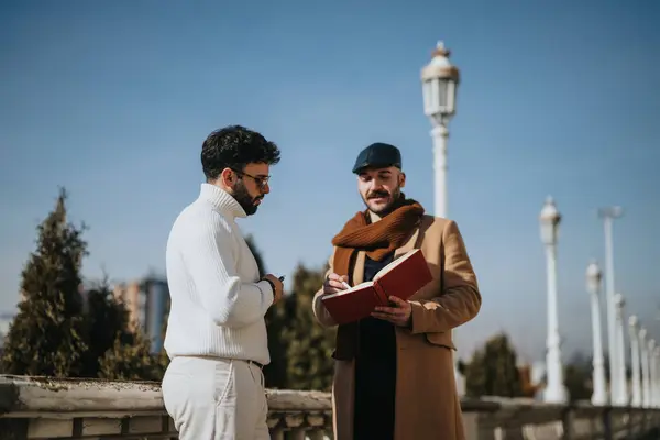 Casual business meeting between two fashionably dressed men, with one holding a notebook, discussing outdoors under clear skies.