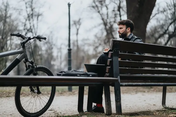 Focused male professional engages in remote work while seated on a park bench, with his bicycle and laptop in an urban outdoor setting.