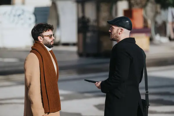 两个穿着时髦的男人在城市人行道上聊天 脸上的表情表明他们正在进行严肃的讨论 明亮的阳光照耀着他们 — 图库照片