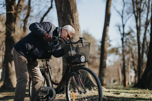Active elderly man pauses during a bike ride to check his bicycle in a tranquil park setting with sunlight filtering through trees.