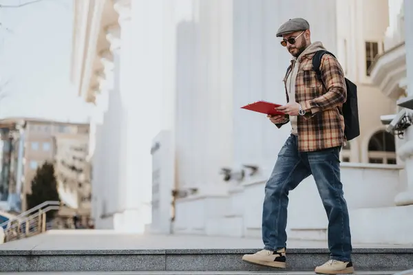 Confident young businessman in casual attire walking outdoors with a clipboard, engaged in work while in a city environment.