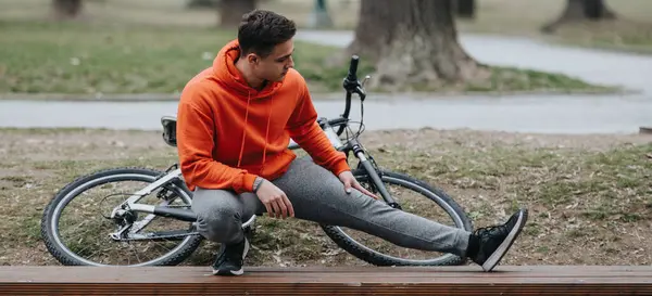 若い大人の男性は自転車の横のベンチでリラックスし 天空の下の屋外公園で休憩を楽しんでいます — ストック写真