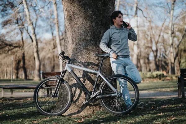 A young man rests against a tree with his bicycle, enjoying a conversation on his smart phone in a serene park setting.