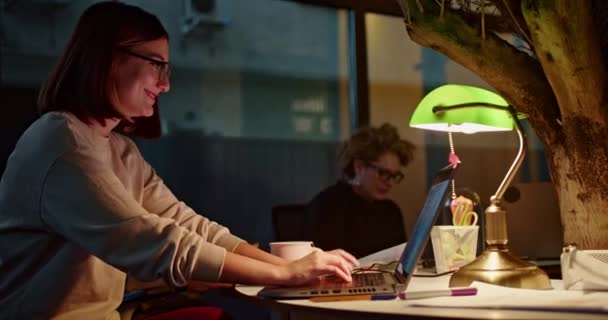 メガネを持つ女性は 暖かく照らされた夜のオフィス環境で彼女のラップトップに集中し 献身と遅い時間の仕事を意味します — ストック動画
