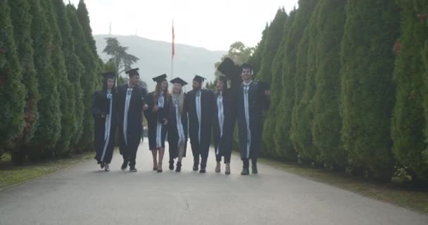 Graduados Jubilosos Universidad Vestidos Con Gorras Vestidos Caminan Parque Exudando — Vídeo de stock