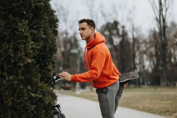オーバーキャストパークの設定で自転車の隣に伸びるために休憩を取るアクティブな若者は 健康的なライフスタイルを示しています — ストック写真