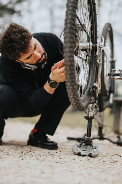 A businessman takes a break from work to focus on fixing the wheel of his bicycle on a gravel path outdoors.