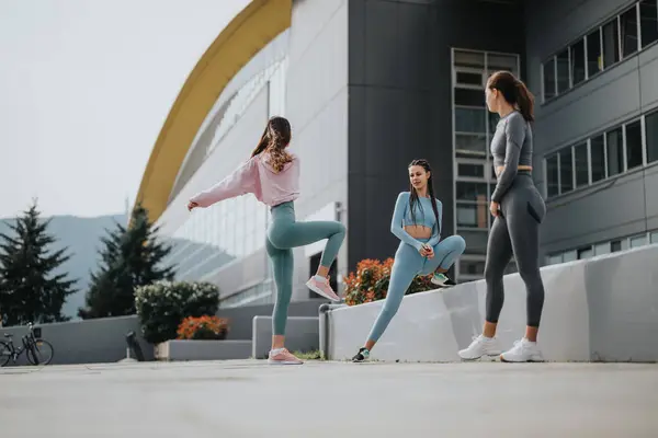 Three women in workout gear preparing for exercise with stretches in an urban setting