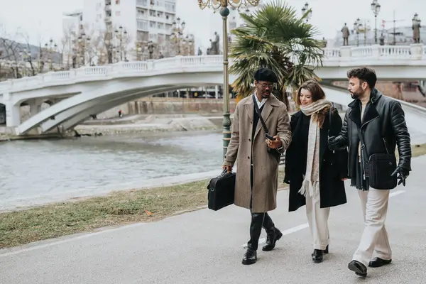 Three fashionable business partners enjoy a walk along a city river promenade, chatting and sharing a moment in their busy urban lives.