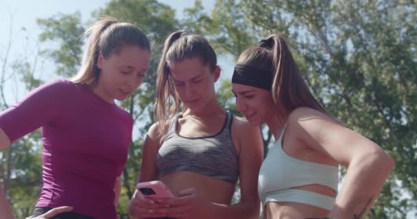 3人のアスレチックな友人は 晴れた公園の設定でスマートフォンを見るために屋外のトレーニングを一時停止します — ストック動画
