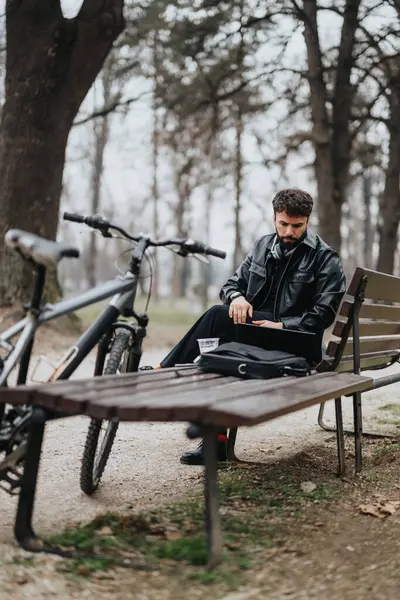 Focused businessman in a leather jacket works on a laptop while sitting on a park bench with his bike beside him, embracing a flexible work lifestyle.