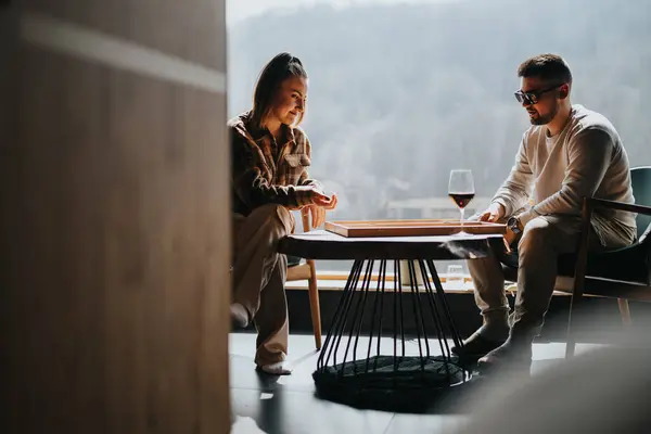 2人の友人は 美しい景色の背景を持つ屋内で ワインのグラスとバックギャモンの古典的なボードゲームを笑い 絆します — ストック写真