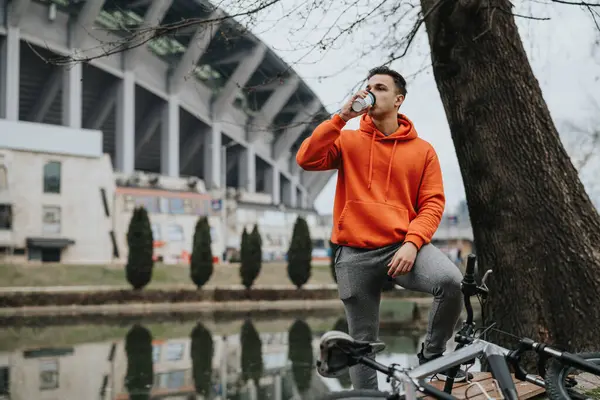 若くてカジュアルな男が静かな都市公園で自転車でベンチでコーヒーを飲んで 曇った空を背景に — ストック写真