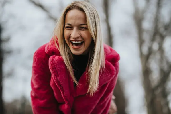 stock image A joyful young blonde woman wearing a striking pink coat is laughing heartily in an outdoor, winter setting.