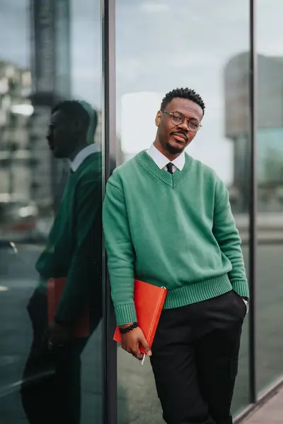 スマートなカジュアルな衣装の若いアフリカ系アメリカ人のビジネスマンは ガラス製のファサードを備えた現代企業の建物の外に自信を持って立っている間 文書フォルダを保持しています — ストック写真