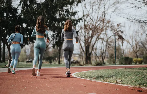 Back view of three female joggers on a running track surrounded by trees, promoting fitness and a healthy lifestyle