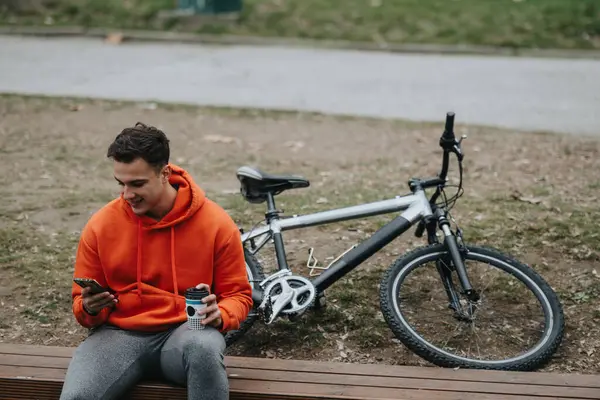 カジュアルな服を着た若い男は 公園のベンチで休んでいる間に携帯をチェックし 彼の隣の自転車で オーバーキャスト日に — ストック写真