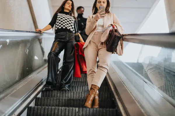 エスカレーターを使用する2人のファッショナブルな女性 1つは彼女のスマートフォンに吸収され 現代の接続性と都市のライフスタイルを示しています — ストック写真