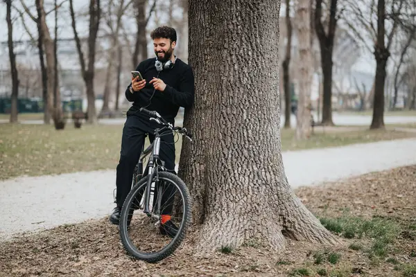 陽気なひげの男は静かな公園の設定で自転車に乗って木の上に休む彼の携帯電話と笑顔をチェックします — ストック写真