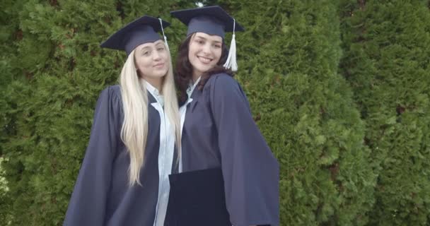 2人の女性卒業生 キャップとガウンのクラッド 誇らしげに彼らのモルタルボードを保持し 緑豊かな背景に対して成功を祝います — ストック動画