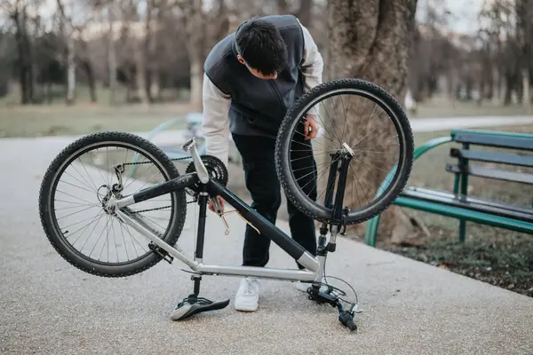stock image Casual male teenager inspects and repairs his bicycle by a bench in a leafy park, representing independence and problem-solving.