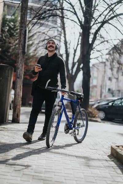 A cheerful man listens to music through headphones while holding a smart phone and standing with his bicycle on a city sidewalk.