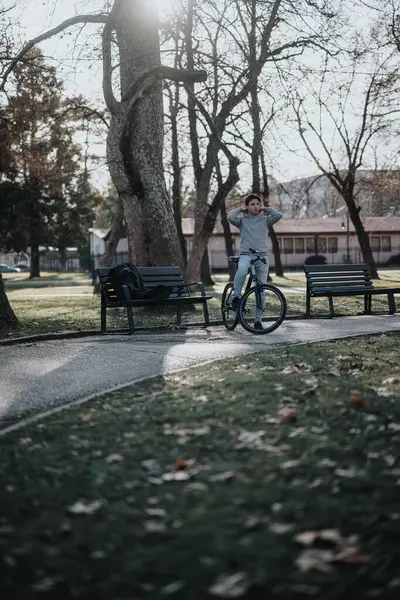 An individual rests with hands behind head on a bicycle in a tranquil park, surrounded by trees and benches.