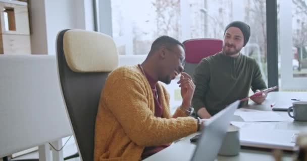 2人の男性従業員 1人のアフリカ系アメリカ人と1人のコーカサス人は 日光に満ちたワークスペースでスピリットな会話に従事し 活気あふれるオフィスの雰囲気を描いています — ストック動画
