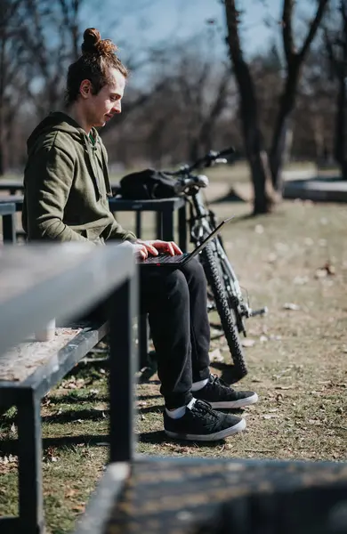 Focused young man with a man bun working remotely on his laptop, seated beside his bike on a park bench on a sunny day.