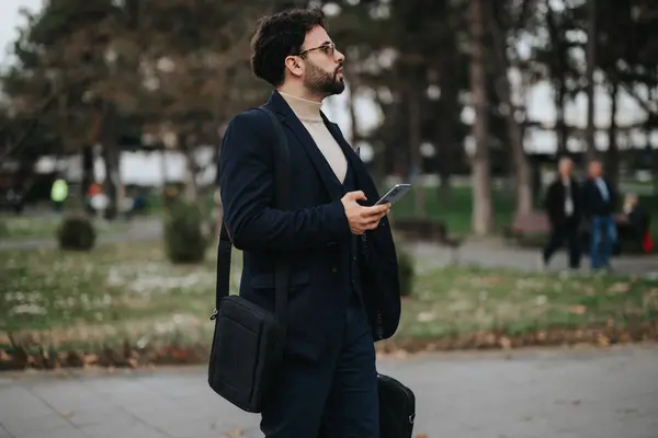 Focused businessman in a suit holding a smart phone and carrying luggage in an urban park setting, possibly awaiting transportation.