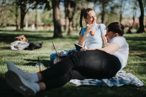 Two young students with textbooks and laptop lounging on a picnic blanket in the park, enjoying a sunny academic day outdoors.