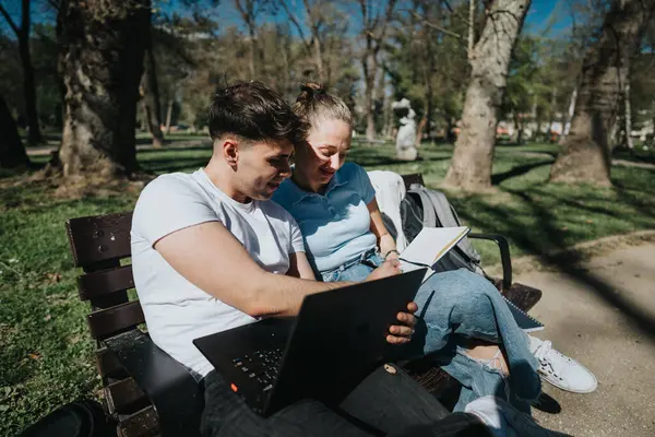 Two friends or students engaged in learning together, utilizing a laptop and notes outdoors surrounded by nature.