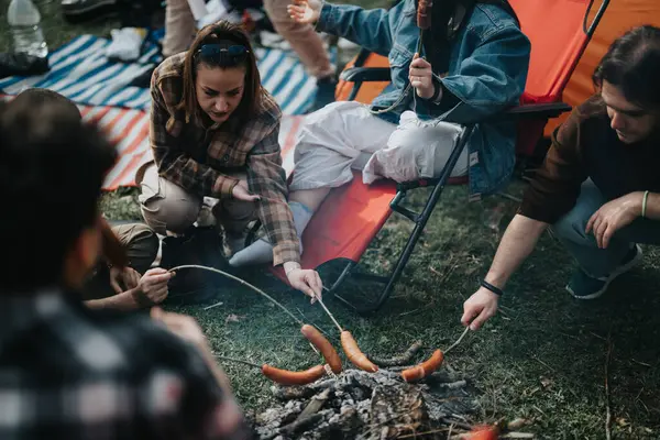 Group of friends gathered around a campfire, roasting sausages and enjoying a laid-back outdoor camping experience.