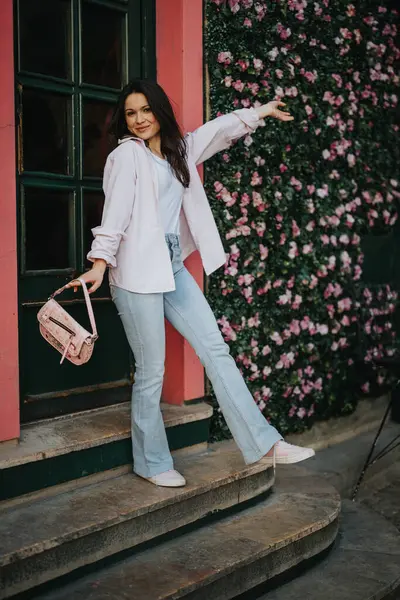 Trendy young woman in light pink shirt and blue jeans with a chic bag standing on steps.